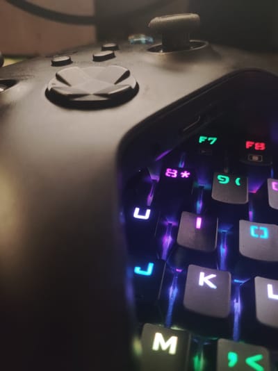 The image is a close-up, low-angle shot of a gaming keyboard and a portion of a game controller. The keyboard features illuminated keys with a variety of colors, including green, red, pink, and blue. Visible keys include "F7", "F8", "*", "9", "(", "U", "I", "K", "L", "M", and what appears to be a comma and angle bracket symbol. The game controller, mostly out of focus, is black and shows some of its buttons and a joystick. The lighting is dim, with the keyboard illumination being the primary light source. There is no indication of the location being Roranwala, Pakistan or any specific urban environment.