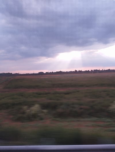 The image depicts a rural landscape at dusk or dawn. A wide, flat field, likely agricultural, dominates the lower half of the frame, showing a blurry motion effect indicative of movement, possibly from a vehicle. The field exhibits variations in color, with patches of green and reddish-brown. A dark, indistinct tree line borders the horizon, punctuated by what appear to be utility poles. Above the field, the sky is filled with heavy, dark clouds tinged with purple and pink hues. A bright shaft of light breaks through the clouds in the upper right portion of the sky. In the foreground, a blurred horizontal element, possibly a railing or barrier, runs along the bottom edge of the image. The general impression is one of a vast, open space under a dramatic sky. The location is specified as Бальшавіцкі сельскі Савет, Belarus.