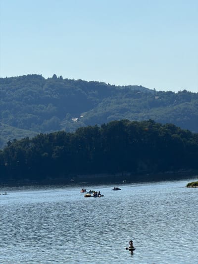 The image depicts a serene summer day at Lake Rożnowskie in Gródek nad Dunajcem, Poland. The clear blue sky forms the backdrop for rolling, tree-covered hills that recede into the distance. In the foreground, the tranquil water of the lake is dotted with various recreational activities. Several small boats, including paddle boats and what appear to be kayaks, are scattered across the surface. A person is visible on a paddleboard in the lower right quadrant of the image, appearing to be holding a paddle. The overall scene conveys a sense of peaceful outdoor recreation on a bright day.
