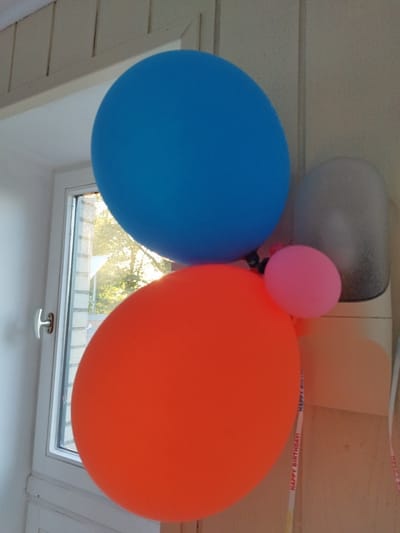 The image shows a close-up of a cluster of balloons. A large, bright blue balloon is positioned at the top. Below it, a large, vibrant orange balloon occupies the lower-left portion of the frame. To the right of the orange balloon and slightly above it, a small, pink balloon is visible. In the background, a gray, translucent balloon or object is partially seen. The balloons are set against a backdrop that includes a window with a white frame and a handle on the left side, through which trees and sunlight are visible, suggesting an outdoor view. The wall appears to be painted white, and there is a wooden paneled ceiling or wall above the balloons. Two decorative ribbons, one white with red text reading "HAPPY BIRTHDAY!", are also present. The scene appears to be indoors, possibly in a domestic setting, and the presence of birthday decorations implies a celebration. The image does not contain any specific landmarks or visual cues that would allow for its precise location within Schiffdorf, Germany.