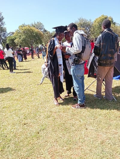 This outdoor photograph depicts a graduation celebration or post-graduation event in Kimumu ward, Kenya, under bright, sunny daylight. The foreground is dominated by a young woman in full graduation attire: a black gown, mortarboard, and a white stole prominently displaying "CLASS OF 2023". She wears silver high-heeled sandals. She, along with another woman partially visible to her left, is looking intently at a mobile phone held by a man standing to her right. This man wears a light grey hoodie, blue jeans, and brown sneakers with white stripes, carrying a backpack and a DSLR camera hanging from his neck. Behind him, another man is seen from the back, dressed in a black leather-like jacket and brown trousers.
In the mid-ground and background, a large number of people are scattered across a expansive grassy field, some standing, others in groups, suggesting a public gathering. Trees line the far background, and a red canopy is visible on the left side. Partially visible behind the camera man is a metal stand or frame, possibly for photography equipment, bearing the brand name "TRiOPO". The overall scene conveys a celebratory and joyful atmosphere, typical of a graduation event.