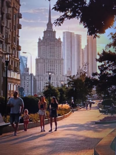 The image depicts a street scene in Balakliya, Ukraine, during what appears to be late afternoon or early evening, given the warm, low sunlight casting long shadows. A prominent Stalinist skyscraper with a spire dominates the background, flanked by modern glass skyscrapers. In the foreground, a paved walkway is lined with trees and planters. Several individuals are walking on the pathway. On the left, a man in a grey t-shirt and shorts is walking with a young child in a pink dress. To their right, two women are walking together; one has long blonde hair and is wearing a black crop top and white shorts, while the other has dark hair and is wearing a dark dress. Further down the walkway, other pedestrians can be seen in the distance. The architecture of the buildings and the overall atmosphere suggest a well-maintained urban environment.