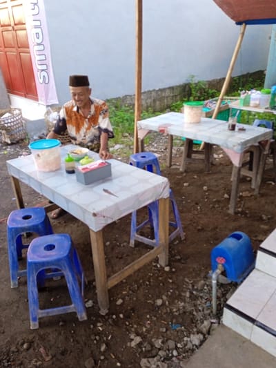 A man wearing a black fez and a patterned shirt sits at a square table covered with a patterned cloth in Pilang, Indonesia. The table holds food items, including a container with a blue lid. Two blue plastic stools sit in front of the table. In the background, another similar table is visible. A blue water container with a faucet stands near a tiled step. The ground appears to be dirt and gravel.