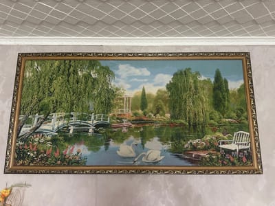 The image shows a framed tapestry depicting a serene garden scene. The tapestry features a tranquil pond with two white swans gracefully swimming on its surface. A white bridge arches over the water in the background, leading to a gazebo nestled amongst lush greenery. Willow trees with cascading branches line the water's edge, their reflections shimmering on the pond. A white bench sits on the bank, offering a spot to admire the picturesque surroundings. The tapestry is framed with an ornate gold-colored border. The tapestry is mounted on a light-colored wall, with a textured white ceiling visible above. No people are present in the scene. The location is Yazykova Pyatina, Russia.