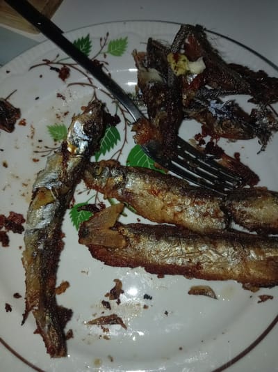 The image displays a close-up, overhead view of a white plate with a decorative rim, featuring a pattern of green leaves and brown branches. The plate contains several fried fish, appearing to be small sprats or similar baitfish, along with scattered remnants of their fried coating and bones. A silver fork is partially visible, positioned diagonally across the plate, with some of the fish pieces appearing to have been disturbed or cut by it. The lighting is dim, casting shadows and highlighting the oily sheen on the fish and plate. The overall scene suggests a meal that has been partially consumed. The location is identified as Тулаўскі сельскі Савет, Belarus.