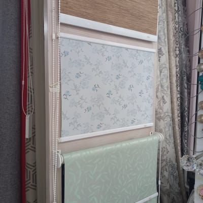The image shows a retail display of window coverings in Briceni, Moldova. In the foreground, a light green roller blind with a subtle leaf pattern is partially unrolled. Above it, a white roller blind with a pale blue floral pattern is displayed. Further up, a textured beige roller blind is visible. To the right of the blinds, several ornate curtains with damask-like patterns in neutral tones are hung. The background reveals more fabric rolls and decorative crystal elements. The display appears to be set up indoors, possibly in a store specializing in home decor or window treatments. There are no people or animals visible in the image.