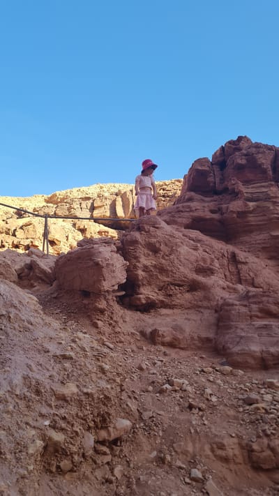 A person stands on a ledge halfway up a reddish-brown rock face against a clear blue sky. The person is wearing light-colored clothing and a red cap. The rock face appears to be part of a cliff or canyon wall, with varying textures and geological formations. A metal railing or support structure is visible along the ledge. The location is in Hevel Eilot Regional Council, Israel.
