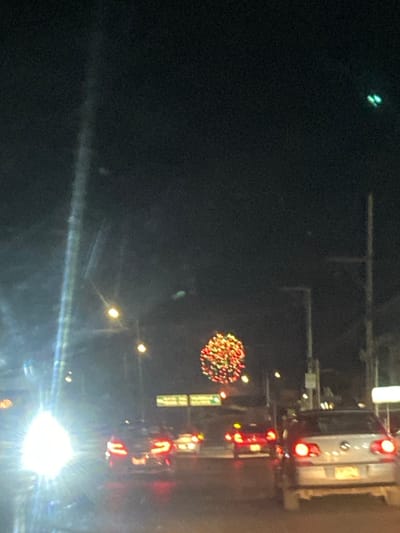 A nighttime street scene features cars with illuminated taillights. Streetlights line the road, and a spherical, colorful decoration hangs above. A sign is visible near the decoration. The image is somewhat blurry, with a streak of light at the bottom, and a dark sky.