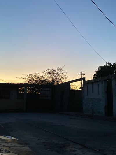 The scene depicts an exterior urban environment in La Cruz del Pastel, Venezuela, during twilight hours (either sunrise or sunset), characterized by low-angle natural light. The sky exhibits a gradient from a pale yellow-orange at the horizon to a deep blue overhead, with a singular faint luminous point visible in the lower-left sky quadrant. The immediate foreground consists of a dark, uneven paved surface, likely asphalt or concrete, with geometric tile patterns partially visible at the bottom-left edge.

The midground is dominated by structures and vegetation silhouetted against the luminous horizon. On the left, a partially constructed or dilapidated building is present, displaying visible concrete columns and a flat roof structure. In the center-right, a dark, incomplete wall or perimeter structure features a distinct rectangular opening that frames the bright horizon. Adjacent to it on the far right, a wall segment with a lighter, textured surface exhibits three vertical rectangular recesses. Sparse deciduous tree silhouettes are interspersed amongst the structures. A utility pole with multiple crossarms and associated wires is silhouetted behind the central wall opening. Overhead utility lines traverse the upper right portion of the frame diagonally. No individuals or specific activities are observable within the depicted scene.