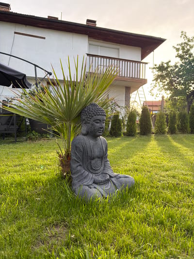 In Lozno, Bulgaria, a gray stone statue of Buddha sits in a meditative pose on a well-maintained grassy lawn. The statue is positioned in front of a palm plant. Behind the statue, a white house with a wooden-gabled roof and a balcony is visible. The sun is setting, casting long shadows across the lawn and creating a warm, golden light. Several coniferous trees are planted in a row along the fence in the background. A black umbrella is partially visible to the left of the statue. The overall scene depicts a peaceful garden setting.
