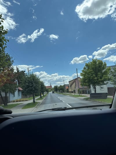 The image shows a view from inside a vehicle looking out at a street in Duszniki, Poland. The street is lined with houses and trees. The sky is blue with scattered white clouds. A church spire is visible in the distance. The road has white lane markings. The vehicle's dashboard and windshield wipers are visible in the foreground.