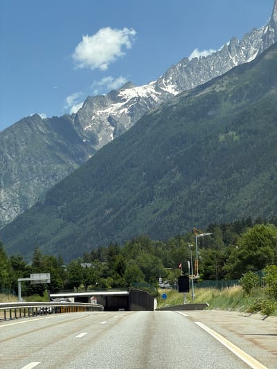 The image shows a highway leading towards a tunnel entrance in Chamonix-Mont-Blanc, France. Towering, snow-capped mountains dominate the background. The highway has white lane markings and guardrails. A paraglider is visible in the sky. In the middle ground, there are trees, buildings, and a construction crane. The sky is blue with a few white clouds.