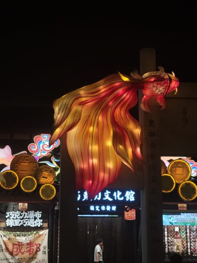 At night, a large, illuminated lantern shaped like a koi fish is displayed in Wuhou District, China. The lantern is primarily red and yellow, with intricate details resembling scales and fins. It hangs suspended, with its head and tail curving dramatically. Below the lantern, signs with Chinese characters are visible, illuminated by neon lights and traditional lanterns. One sign in blue neon reads "文化館" (Culture Center). In the foreground, a person in a light-colored shirt is walking, and another person's back is partially visible. Several large, circular coin-like lanterns are also lit up, adding to the festive atmosphere. The background is dark, suggesting it is nighttime.