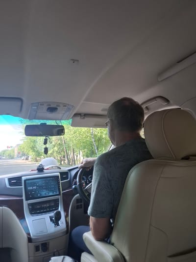 An interior view of a car in Svobodny, Russia, shows a man driving. He has graying hair and is wearing a light blue t-shirt with a palm tree pattern. The car's dashboard features a touchscreen infotainment system displaying radio frequencies, and a gear shift is visible. A rearview mirror hangs from the ceiling, adorned with two decorative items. Outside the car, trees line a street with buildings and traffic lights in the distance. The passenger seat in the foreground is a light beige leather.