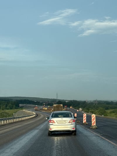 A silver Toyota Corolla is traveling on a wet highway. The car has a license plate that reads "X504OP 196". The highway appears to be undergoing construction, with orange and white traffic cones and barriers visible. In the distance, there are trees and buildings, and a tall structure resembling a communication tower. The sky is overcast with some clouds. The image is taken from a slightly elevated perspective, looking forward from behind the vehicle. The location is identified as the city of Boyarka, Russia.
