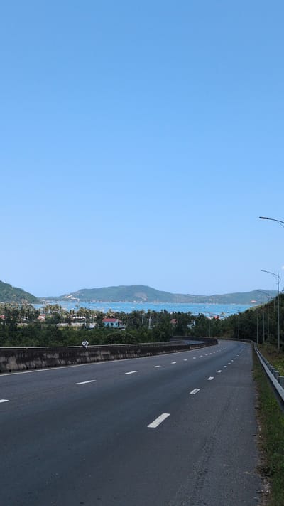 The scene depicts an elevated asphalt highway in Hòa Hiệp, Vietnam, under a clear, cloudless blue sky. The road, marked with white dashed lines, curves gently to the right and ascends. A grey concrete barrier lines the left side of the road. Below this barrier, a densely vegetated area featuring numerous trees and palm trees contains several residential structures with tiled roofs, including one prominent light blue house and another with a reddish-brown roof. A small, light-colored figure, appearing to be a person, is seated on the concrete barrier, facing away from the road towards the bay. The right side of the highway is bordered by a metal guardrail and a vegetated embankment, with two dark streetlights visible. Beyond the immediate terrain, a wide bay is present, its blue water dotted with numerous small boats or aquaculture installations. The far horizon is defined by multiple green, tree-covered hills or islands rising from the bay.
