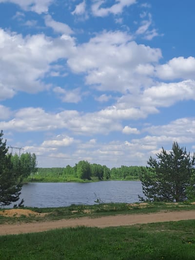The image depicts a body of water bordered by greenery on one side and a grassy area with a dirt path on the other. The sky is blue with numerous white clouds. Power lines are visible in the distance. The location is Avdot'ino, Russia.