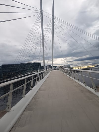 The image depicts a modern pedestrian bridge in Drammen, Norway, under a cloudy sky. The bridge features a white railing and a concrete walkway that recedes into the distance. Tall, white, mast-like structures rise from the bridge, supporting numerous thin cables that fan out to hold the walkway. To the left, a large, dark, modern building with many windows is visible, reflecting the overcast sky. Further in the distance, across a body of water, other buildings and another bridge are discernible. The overall impression is one of an urban landscape on a gloomy day, possibly late afternoon or early evening, given the subdued light and the hint of warmer tones on the horizon in the distance. There are no people visible, and the scene is static, focusing on the architecture of the bridge and its surroundings. The weather appears to be overcast with some texture in the clouds, suggesting a mild or cool day.