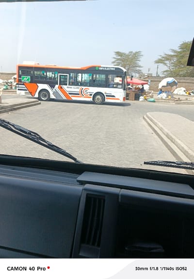 The image captures an outdoor scene from the perspective of a vehicle's interior, looking through the windshield. The dark dashboard and windshield wipers are visible in the foreground, indicating the viewpoint.
The main subject is a modern-looking bus, which dominates the middle ground. The bus is predominantly white with striking orange, grey, and black geometric patterns along its sides. Text on the bus clearly reads "REMBO CLASSIC" and a large "S" within a circular logo. Towards the rear top of the bus, the phrase "100% ELECTRIC" is visible, accompanied by a green leaf icon, indicating its environmentally friendly nature. The bus appears to be stationary, parked on a paved area, possibly a bus stop or roadside.
In the background, to the right of the bus, there are several makeshift stalls or vendor setups, some covered with red and white umbrellas, suggesting a market or street vendor scene. People are faintly visible near these stalls, appearing to be engaged in daily activities. Further beyond, sparse trees with light green foliage are visible, and some low-rise structures or buildings line the horizon. The ground around the bus and stalls is paved with interlocking bricks, while further back, it seems more uneven. The sky is bright and clear, indicating a sunny daytime setting.
Technical information embedded in the image indicates it was captured with a "CAMON 40 Pro" camera, with settings of "30mm f/1.8 1/1140s ISO52".