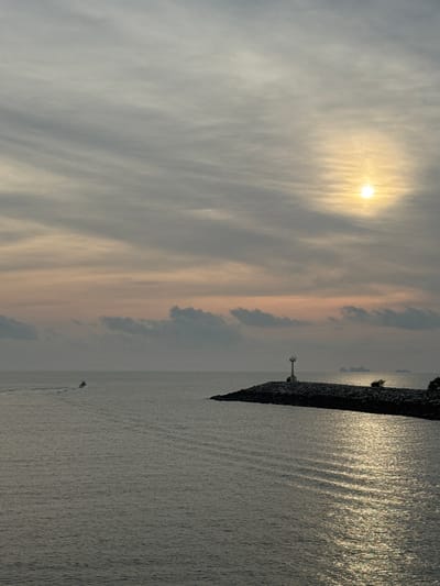 The image shows a coastal scene in Pak Nam Chumphon, Thailand, taken at either sunrise or sunset, as evidenced by the low-angle sunlight piercing through the clouds. A breakwater extending into the sea is visible on the right, with a small lighthouse or navigational aid structure at its tip. The water surface is textured with wavelets and reflections of the sun. A small boat can be seen moving on the water's surface in the distance. The sky is covered with a mix of clouds.