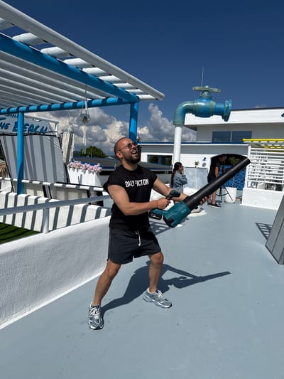 In Baliovtsi, Bulgaria, a man in a black t-shirt and shorts holds a leaf blower, aiming it upwards. He wears sunglasses and appears to be laughing or shouting. He is on a grey, outdoor surface, possibly a rooftop or deck, with white railings and a blue structure overhead. In the background, a large, blue, decorative faucet is mounted on a white pole. Other people and structures are visible further in the background, including a white building with windows and a yellow railing. The sky is blue with scattered clouds.