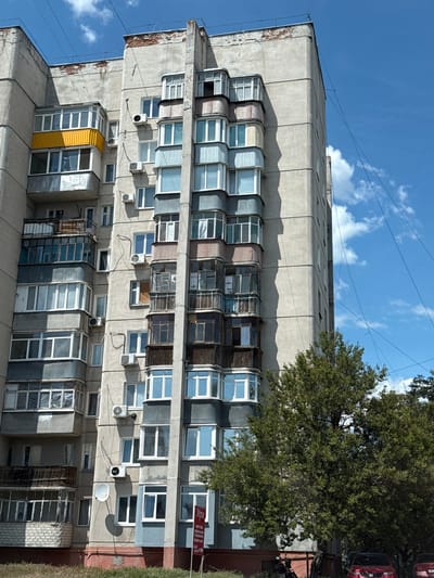 A tall, multi-story residential building, likely an apartment block, dominates the scene, characterized by its concrete facade. The building features numerous windows and enclosed balconies (loggias) that vary significantly in design, materials, and condition, indicating individual modifications by residents. Many external air conditioning units are visible on the facade. A large, leafy green tree partially obscures the right side of the building, and the roofs of a few red cars are faintly visible behind it. Overhead, wires or cables stretch across a bright blue sky with scattered white clouds. A white satellite dish is attached to one of the lower balconies.
No specific activity or event is depicted; the image provides a static view of the building.
The photograph was taken during the daytime, evidenced by the bright sky and strong shadows cast by the building's overhang and balconies. The architectural style, with its gray concrete and repetitive structure, suggests a typical Soviet-era residential building found in Eastern Europe, consistent with the location context of Balakliya, Ukraine. The building shows signs of age and wear, including discolored concrete and some exposed material on its corners. The diverse range of balcony enclosures, from modern glass to older, weathered structures, including a distinctive bright yellow enclosed balcony, adds to the building's lived-in appearance.
A red and white sign, partially hidden by the tree on the right side of the building, displays the Cyrillic letters "MYPC" (likely "МИР", meaning "Peace" or "World") along with an arrow pointing to the right.