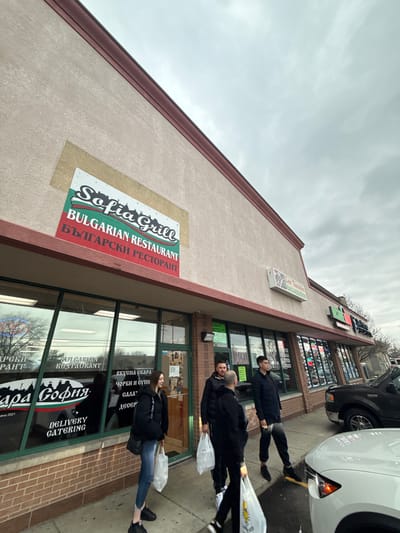 The image shows the Sofia Grill, a Bulgarian restaurant located in Des Plaines, United States of America. The restaurant has a sign in English and Bulgarian, stating "Bulgarian Restaurant" and "БЪЛГАРСКИ РЕСТОРАНТ." The establishment date is 2021. The building appears to be a strip mall with multiple storefronts. There are people walking on the sidewalk in front of the restaurant, some carrying bags. Cars are parked along the curb. The weather is overcast.