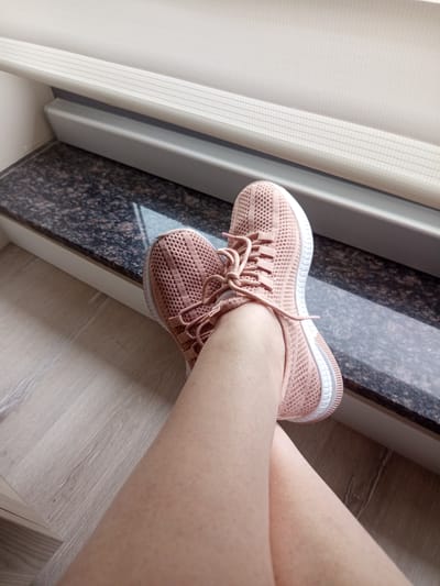 A close-up, over-the-shoulder shot shows a person's legs wearing pink knitted athletic shoes. The shoes are laced with pink laces. The person is sitting on a floor with a light wood grain pattern, next to a dark granite countertop. The countertop is located near a window with a white blind. The scene appears to be indoors, likely in a residential setting. There is no indication of the specific location within Ankara, Turkey.