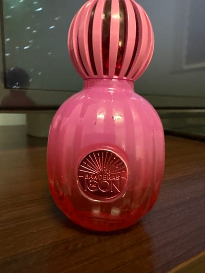 A close-up, eye-level, slightly upward-angled shot shows a pink, ribbed, globe-shaped perfume bottle with a red interior. The bottle has a round, metallic emblem on the front with the inscription "- BANDERAS ICON -". The bottle sits on a dark wooden surface. In the background, out of focus, is a dark screen, possibly a television, with some white specks and a faint reflection. There are no people visible in the image. The location in Małkowo, Poland, is not discernible from the image.