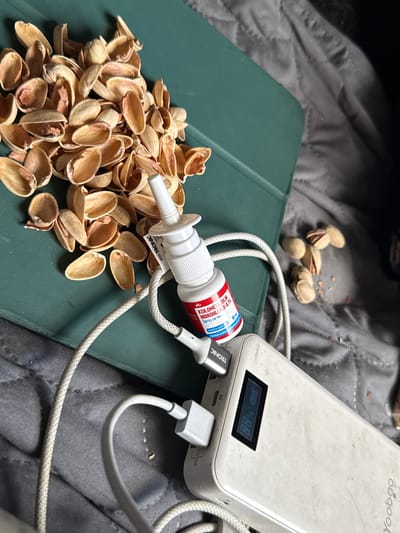 This close-up, medium-shot image depicts a collection of pistachio shells scattered on a dark green, tablet-like surface. The shells are light brown and vary in size and shape, with some appearing more broken than others. A small, white nasal spray bottle with a red and blue label is positioned amongst the shells, its nozzle pointing towards the left. A white cord is attached to the spray bottle and runs towards a white power bank, which is also partially visible in the frame. The power bank has a digital display showing "88%" and a battery icon. Another white cord is plugged into the power bank. The background is a dark gray, quilted fabric, possibly a blanket or bedspread. A few whole pistachios are visible further up and to the left of the frame. The lighting appears to be artificial and casts shadows. The scene is set indoors, as indicated by the power bank and the fabric background. There are no people visible in the image. The overall impression is one of a still life arrangement, possibly suggesting a moment of rest or break, with the nasal spray and charging device hinting at personal use. The brand "TRONIC" is visible on the power bank and the spray bottle's label. Text on the spray bottle is partially legible, including "KSILOMETAZOLIN HIDROHLORID 0,1% Sprej za nos". The location context is Kanjoši, Montenegro.