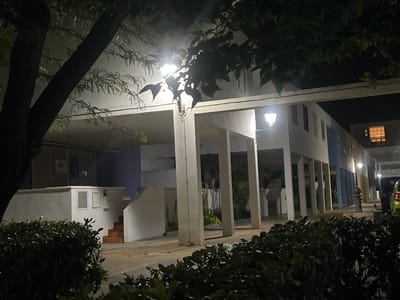 This is a nighttime photograph taken from ground level, looking towards a building complex in Alcàsser, Spain. The scene is illuminated by streetlights and ambient light from within the buildings, casting long shadows and creating a somewhat ethereal atmosphere.

In the foreground, dark green, leafy bushes form a dense hedge, obscuring the lower portion of the image. To the left, the dark trunk and branches of a large tree are prominently featured, with its leaves catching the light from above and appearing somewhat wet or dewy.

The main subject of the image is a multi-story building with a distinctive architectural style. It features a series of white, rectangular columns supporting an upper level or covered walkway. The building's facade is painted in muted tones, with sections of white, light blue, and yellow visible. There are visible windows with louvre-style shutters and some windows illuminated from within, particularly on the upper floors.

A staircase with orange treads leads up the side of a white structure on the left, adjacent to the building. A white utility box or access panel is mounted on this structure. Further to the right, a streetlamp with a white, hexagonal shade is mounted on an arm extending from the building.

In the background, the outline of other buildings can be seen, and a bright yellow window with multiple panes is visible on the right side of the frame. A portion of a green vehicle is parked on the right side of the image, partially obscured by the bushes.

The overall environment appears to be a residential or communal outdoor area within a town or city. The presence of the tree, bushes, and streetlights suggests a public space. The lighting conditions indicate it is after dark. There are no people visible in the photograph.