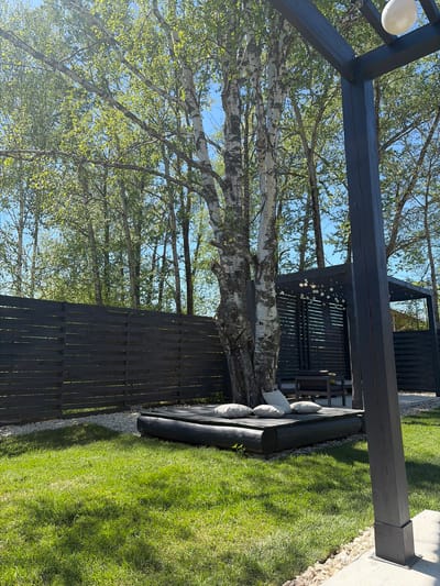 A slightly rotated medium shot shows a lawn with a black pergola in the city of Юг, Russia. The yard has a short green lawn. A wooden pergola structure is in the top left of the shot, with a white light fixture hanging from the corner. A pale, spotted tree trunk is in the center-left. In the background, there is a structure with a wooden lattice design. The lawn has a seating area with white pillows on black cushions. Wooden lattice fencing is around the perimeter of the lawn.