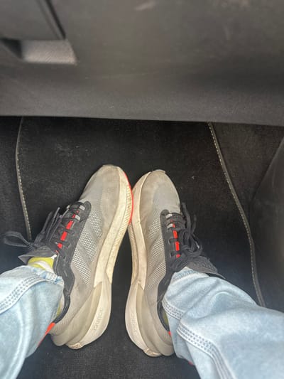 The image captures a downward, first-person perspective of an individual's feet and lower legs within a vehicle interior, located in Torroella de Montgrí, Spain. The subject is wearing light blue denim jeans, yellow socks with white horizontal stripes, and a pair of grey athletic shoes. The shoes feature black lacing systems with three red eyelet loops on each shoe, a mesh upper with a distinct pattern of small white dots forming three stripes, and thick off-white soles marked with the word "BOUNCE." The footwear shows visible dirt and scuff marks, particularly on the soles and lower uppers. The feet rest on a black carpeted floor mat, with a dark grey, textured plastic or fabric surface visible directly above it, consistent with a vehicle's dashboard or lower console area. No other persons or specific actions are depicted.