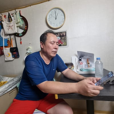 A man is sitting at a table, looking at a smartphone. He is wearing a blue t-shirt and red shorts. The room is cluttered, with bags and pictures hanging on the wall. A clock on the wall shows the time is 1:05. There are also bottles of hand sanitizer and water on the table.