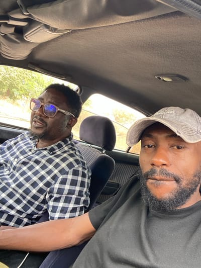Two adult males are depicted inside a vehicle, consistent with a passenger car, in Ungwan Liman, Nigeria. The image is captured from the perspective of the male in the foreground, positioned on the right. This individual, estimated 30-40 years old, has a dark complexion, a short dark beard and mustache, and wears a black t-shirt and a grey, distressed baseball cap with faint white text. He looks directly at the camera with a neutral expression. To his left, partially in the background, sits another male, estimated 40-50 years old, with a dark complexion, short dark hair, a beard, and square-framed glasses with light-colored lenses. He wears a blue and white checkered long-sleeved shirt and is looking towards the left, away from the camera, with his mouth slightly open. Both individuals are seated in dark-colored car seats featuring a patterned fabric on the backrests. The vehicle's interior includes a light grey fabric headliner. Through the car windows, green foliage and trees are visible under daylight conditions.