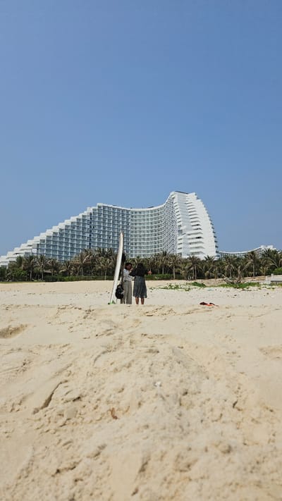 The image shows a beach scene, with a large white hotel structure in the background and blue skies. Two people stand with a surfboard on the sand. The location is Bac Cam Ranh, Vietnam. The beach sand is light-colored, and palm trees are visible between the hotel and the beach.