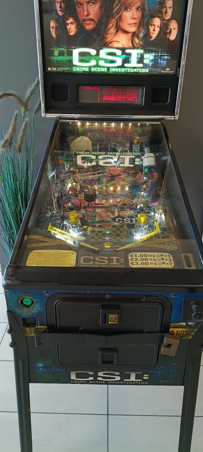 This is a vertical shot of a CSI: Crime Scene Investigation pinball machine. The machine is dark blue with graphics of the CSI cast and the CSI logo. The playfield is visible under the glass, filled with ramps, targets, and flashing lights. A red LED display on the side shows "PINBALLINGE" and some numbers. The machine has a start button and what appears to be a coin slot. The background suggests an indoor setting, possibly a game room or arcade, with a tiled floor. The overall impression is that the pinball machine is not currently in use.