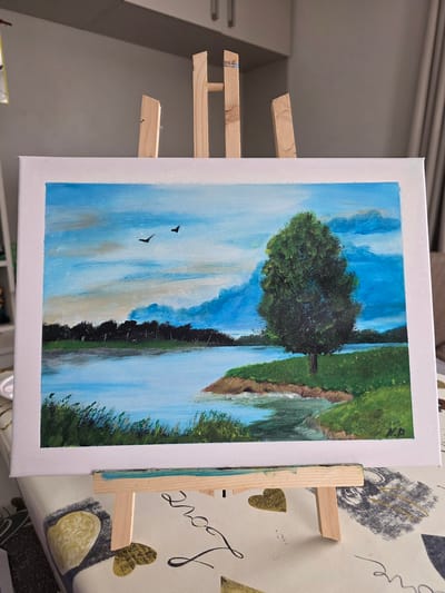 This media file displays a painting on a canvas, which is mounted on a wooden easel. The painting depicts a scenic landscape with a prominent tree on the left, a body of water, and a grassy bank on the right. The sky is a vibrant blue with some clouds. Two small, dark bird silhouettes are visible flying in the sky. The scene appears to be set during the daytime, with bright lighting suggestive of good weather. The style of the painting is impressionistic, with visible brushstrokes. The canvas is white-bordered. The easel itself is made of light-colored wood and is placed on a surface covered with a patterned cloth, which includes floral motifs and the word "love" written in cursive. There are no people visible in the painting or the surrounding environment. The overall impression is one of a peaceful, natural setting. The location context is provided as Claremorris, Ireland, though there are no specific visual cues within the painting to confirm this location.