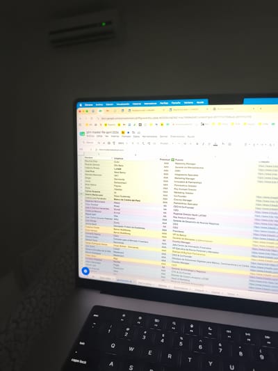 Here is a detailed description of the image:
**Overall Scene:** The image shows a laptop screen displaying a Google Sheets spreadsheet titled "gtm master file april 2026." The laptop is placed on a surface, and the background is dark, with what appears to be an air conditioner visible in the upper part of the frame.
**Laptop Screen Content:** The spreadsheet appears to be a database or list of contacts, likely for a go-to-market (GTM) strategy. The columns include:
- Nombre (Name)
- Empresa (Company)
- Prioridad (Priority)
- Puesto (Job Title)
- LinkedIn (with corresponding URLs)
The data in the sheet includes names, company affiliations, priority levels (primarily "AAA" and "AA"), job titles (e.g., "Marketing Manager," "CEO," "Country Manager"), and LinkedIn profile links. There are several companies listed, representing a variety of industries like fintech, banking, and more.
**Location:** Given the "guatemala 26" and "haz tu pizza" text in the sheet tabs, the contacts being managed may be for Guatemala.
The row labeled "Valeria Maldonado" which also has "Nequi Guatemala" in the Empresa (Company) column, this places the location in Guatemala, specifically in Guatemala City, Guatemala, as "Nequi" is a well-known brand in the city.
**Laptop and Keyboard:** The laptop is a dark color with a black keyboard. Keys such as 'esc,' 'tab,' 'Q,' 'W,' 'E,' 'R,' 'T,' 'Y,' 'U,' 'A,' 'S,' etc., are visible. The keyboard appears modern.
**Environment:** The background is dim, suggesting a low-light setting.