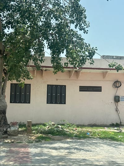A medium shot captures a pale pink building with a red-trimmed roof. Two dark-framed windows with white bars are visible on the left, followed by a smaller, dark-framed horizontal window. To the right, an electrical meter box is mounted on the wall, with coiled black wire above it. The building is partially obscured by the lush green foliage of a large tree in the foreground on the left. The ground in front of the building is a mix of green vegetation, scattered debris including plastic bags, and a paved area with interlocking bricks. A wooden post with carved markings stands near the tree. The scene is set against a clear blue sky, suggesting an outdoor environment in Ajmer, India.