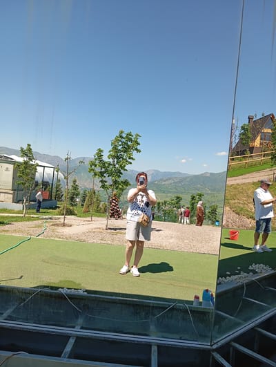 The image shows a woman taking a photo with a smartphone, likely a self-portrait, in Chinarkent, Uzbekistan. She is standing on a green artificial turf area, part of an outdoor recreation space with trees and a view of distant mountains under a clear blue sky. A building with a white dome is visible in the background. In the foreground, a glass structure reflects the scene, including a house and a man standing nearby. Other people are also visible in the background.