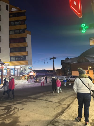 This image depicts an illuminated street scene at dusk in Saint-Martin-de-Belleville, France, characterized by a mix of pedestrian activity and alpine architecture. The sky presents a gradient from deep blue overhead to a lighter orange hue at the horizon.
In the immediate foreground, facing away from the camera and towards the midground, stands an individual wearing a white fleece jacket, dark blue jeans, a grey knitted beanie with a pom-pom, and a dark shoulder strap across their back. The paved street surface, with visible snow patches, occupies the lower third of the frame. A circular manhole cover is present in the lower-left quadrant.
To the left, a multi-story building with a white facade and horizontal light-brown wooden balcony railings rises prominently. Snow is visible on its roof. The ground floor houses illuminated storefronts, including one with "RENS ALBIER" visible and a small, brightly lit wooden chalet-style kiosk with snow on its roof, displaying a sign that reads "PLAT DU JOUR". Several individuals are walking in this area; two in the left foreground include a male in a red jacket and a female in a grey jacket.
In the midground, multiple pedestrians are observed walking. A group of three individuals, including one in a vibrant pink jacket, and another person in a blue and white jacket, are present. A white object, possibly a small animal or sculpture, is situated near a white van parked on the street. A prominent ski lift pylon with cables and two empty chairs extends vertically against the sky. Other snow-covered buildings are visible in the distance.
Overhead, to the right, a bright red, diamond-shaped neon sign is partially visible. Below it, a green illuminated cross sign, characteristic of a pharmacy, glows, with partial text "OUV" visible. A yellow sign bearing "sherpa" is also partially visible. The background reveals distant snow-covered mountains silhouetted against the twilight horizon.
