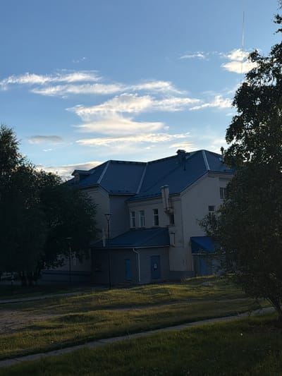 In the city of Kandalaksha, Russia, on a clear day with scattered clouds, a light-colored, multi-story building with a blue roof is depicted. The building has multiple windows and appears to be surrounded by trees. A grassy area with a path in the foreground suggests an outdoor setting, possibly a park or residential area. The lighting indicates it is daytime.