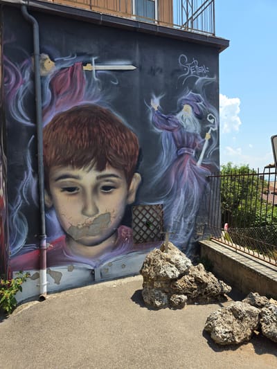 This media file depicts a large mural painted on the exterior wall of a building, likely in Viterbo, Italy. The mural is oriented vertically. The primary subjects are a stylized depiction of a young boy's head and a mystical figure. The boy's face is rendered with detailed shading, showing hints of cracking paint on his forehead, and his eyes are open but appear vacant. He has brown hair. Above and behind him, a figure in flowing purple robes with long white hair and a beard is depicted. This figure holds a glowing staff or wand and appears to be conjuring ethereal wisps of smoke or energy. A sword, with a golden hilt and a silver blade, is positioned vertically to the left of the boy's head, seemingly emerging from a swirl of red and purple fabric.
The setting appears to be an urban environment, with a building facade visible to the left, featuring a window and a downspout. The sky is bright blue with scattered white clouds, suggesting it is daytime, likely a sunny day. To the right of the mural, a rough, textured concrete surface slopes upwards, with large, irregularly shaped rocks placed on it. A metal fence with vertical bars and a horizontal railing runs along the top of the wall, behind which green foliage is visible. A portion of a metal scaffolding structure is visible on the far left of the image, extending upwards.
The overall mood of the mural is somewhat enigmatic and fantastical, with the juxtaposition of the boy's realistic yet somewhat haunting portrayal and the magical elements. There is some visible text in the upper left quadrant of the mural, which appears to be stylized calligraphy or graffiti, possibly in an Asian script.