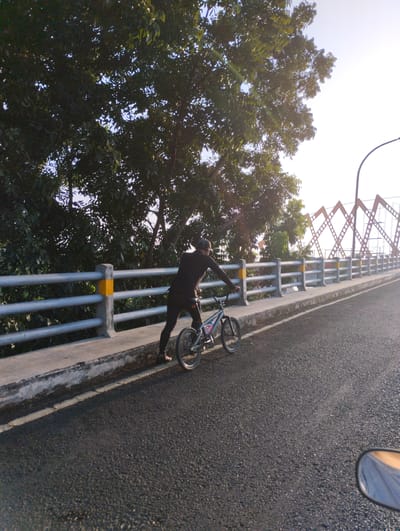 In Sidodadi, Indonesia, a person dressed in black is on a BMX bicycle on a bridge with a metal railing. The cyclist is facing away from the camera and appears to be pushing the bicycle. The bridge has a concrete walkway with a yellow stripe on the railing. Trees with dense foliage are visible behind the railing, and the sky is bright and clear with the sun setting. In the foreground, a motorcycle mirror is visible, reflecting a person wearing a blue shirt.