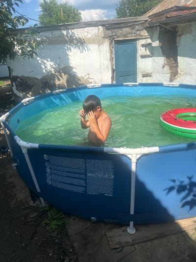 The image shows a young boy in an inflatable swimming pool filled with green water. He is wearing swimming goggles and appears to be adjusting them. The pool is set up in a yard with a dilapidated building in the background, characterized by peeling paint and a weathered appearance. A blue wooden door is visible on the building. A red and green inflatable ring, resembling a watermelon, floats in the pool. The ground around the pool is dirt and debris. The overall scene suggests a summer day in a rural or semi-urban setting.