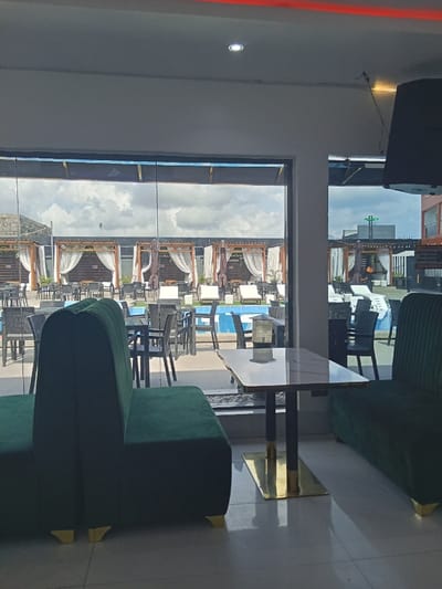 The image shows the interior of a cafe with a large window looking out onto an outdoor area with a swimming pool. The cafe is furnished with two large green couches with gold bases, a black and gold square table, and several black chairs. The window is large and reveals a bright outdoor area with lounge chairs, umbrellas, and a swimming pool. The sky is partly cloudy. There are no people present. The scene is in Ujevwu, Nigeria, at coordinates latitude: 5.471, longitude: 5.818.