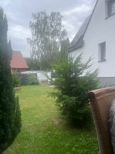 The image depicts a backyard scene in Garbsen, Germany, on an overcast day. A white house with solar panels on its roof is visible on the right side of the frame. Adjacent to the house, a section of a red-tiled roof belonging to another building is visible on the left. The yard is covered in green grass and various types of trees and bushes. In the foreground, a portion of a brown wicker chair is visible. The background shows more trees and a fence. The overall impression is that of a residential area with well-maintained greenery.