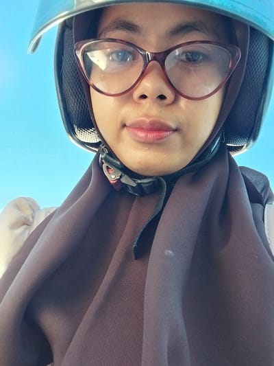 A close-up, low-angle, selfie-style photograph captures a person wearing a blue helmet and a brown hijab. The person has on red-rimmed cat-eye glasses. The background is a clear blue sky. The scene appears to be outdoors. The individual's face is the central focus.