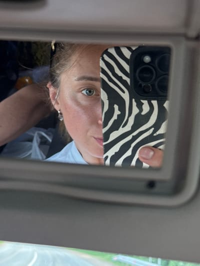 A woman is taking a selfie in a car using the visor mirror. She is holding a cell phone with a zebra-striped case. The car's interior is visible, with the mirror surrounded by a gray frame. The environment outside the car is blurred and green, possibly trees or foliage. The location is not discernable from the image.